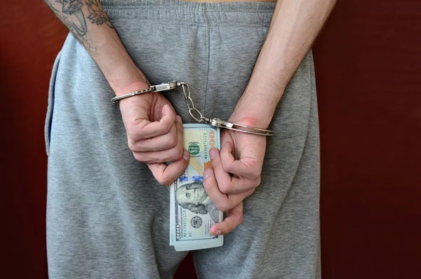 An arrested guy in gray pants with handcuffed hands holds a huge amount of dollar bills. Back view.
