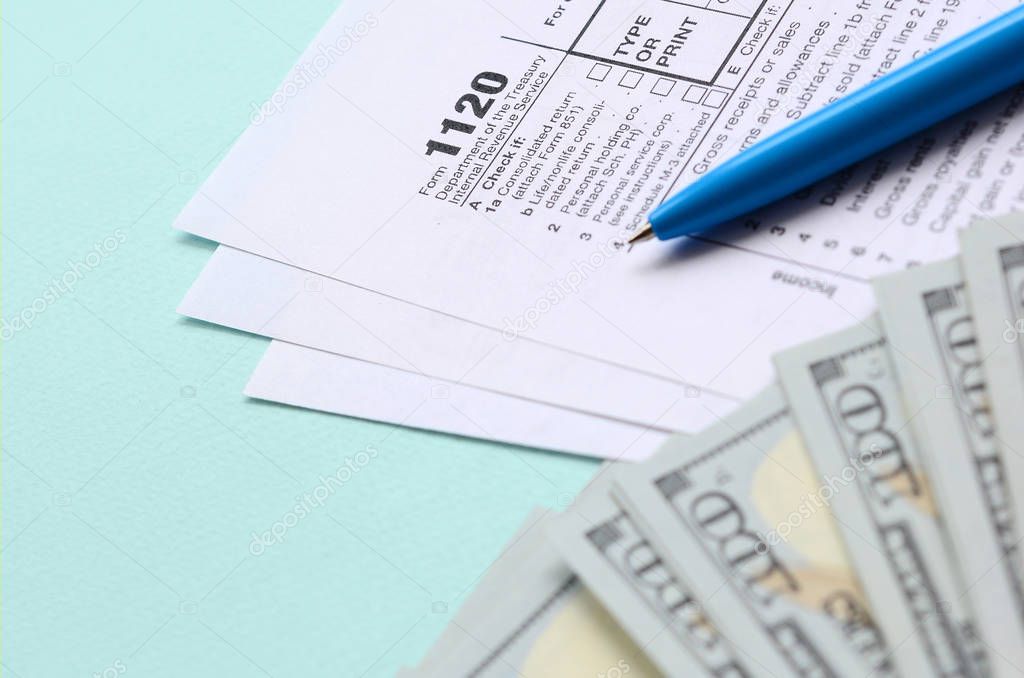 1120 tax form lies near hundred dollar bills and blue pen on a light blue background. US Corporation income tax return.