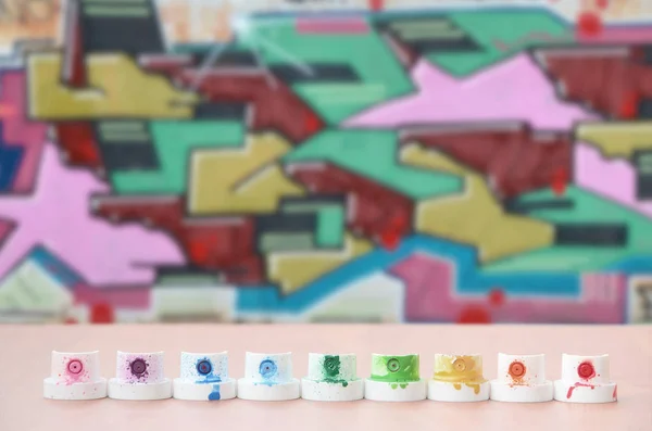 The soiled multicolored nozzles from the paint sprayer are lined up on a wooden table on a background of colored graffiti drawing in a wild style. The concept of street art