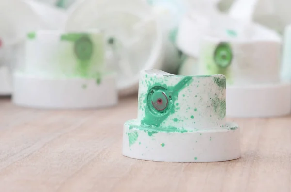 Several plastic nozzles from a paint sprayer that lie on a wooden surface against a gray wall background. The caps are smeared in green paint. The concept of street art and graffiti