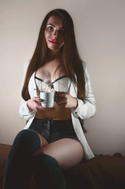 Genç ve seksi beyaz esmer kız bir beyaz gömlek ve beyaz iç çamaşırı portresi beyaz fincan sıcak kahve bej duvarlı bir odada oturuyor ve ellerde ile.