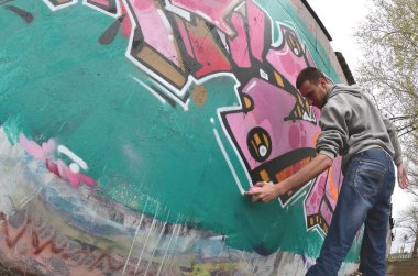 Gri kapüşonlu genç bir adam, yağmurlu havalarda grafiti bir duvar pembe ve yeşil renklerde boyar. Balıkgözü atış