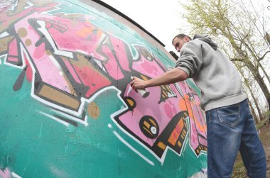 Gri kapüşonlu genç bir adam, yağmurlu havalarda grafiti bir duvar pembe ve yeşil renklerde boyar. Balıkgözü atış