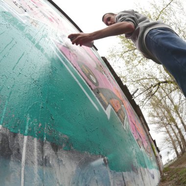 Gri kapüşonlu genç bir adam, yağmurlu havalarda grafiti bir duvar pembe ve yeşil renklerde boyar. Balıkgözü atış