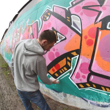 Gri kapüşonlu genç bir adam, yağmurlu havalarda grafiti bir duvar pembe ve yeşil renklerde boyar. Balıkgözü atış