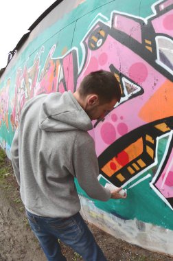 Gri kapüşonlu genç bir adam, yağmurlu havalarda grafiti bir duvar pembe ve yeşil renklerde boyar. Balıkgözü atış