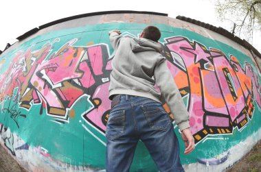 Gri kapüşonlu genç bir adam, yağmurlu havalarda grafiti bir duvar pembe ve yeşil renklerde boyar. Balıkgözü atış