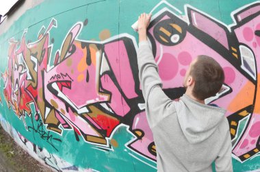 Gri kapüşonlu genç bir adam, yağmurlu havalarda grafiti bir duvar pembe ve yeşil renklerde boyar. Balıkgözü atış
