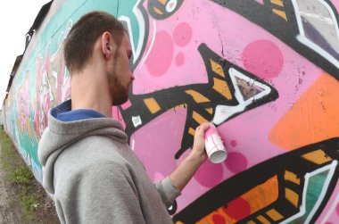 Gri kapüşonlu genç bir adam, yağmurlu havalarda grafiti bir duvar pembe ve yeşil renklerde boyar. Balıkgözü atış