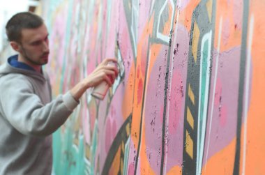 Gri kapüşonlu genç bir adam, yağmurlu havalarda grafiti bir duvar pembe ve yeşil renklerde boyar. Duvar ve bulanık sanatçı parçası odaklanmak