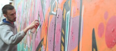 Gri kapüşonlu genç bir adam, yağmurlu havalarda grafiti bir duvar pembe ve yeşil renklerde boyar. Duvar ve bulanık sanatçı parçası odaklanmak