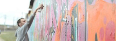 Gri kapüşonlu genç bir adam, yağmurlu havalarda grafiti bir duvar pembe ve yeşil renklerde boyar. Duvar ve bulanık sanatçı parçası odaklanmak