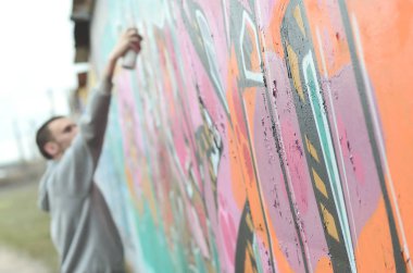 Gri kapüşonlu genç bir adam, yağmurlu havalarda grafiti bir duvar pembe ve yeşil renklerde boyar. Duvar ve bulanık sanatçı parçası odaklanmak
