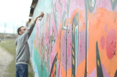 Gri kapüşonlu genç bir adam, yağmurlu havalarda grafiti bir duvar pembe ve yeşil renklerde boyar. Duvar ve bulanık sanatçı parçası odaklanmak