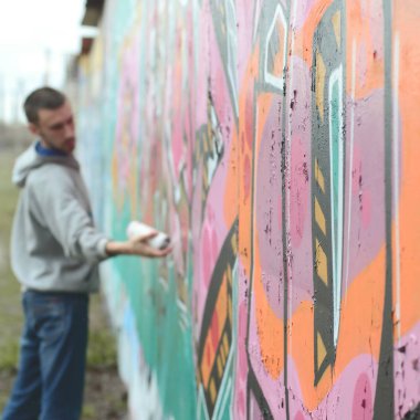 Gri kapüşonlu genç bir adam, yağmurlu havalarda grafiti bir duvar pembe ve yeşil renklerde boyar. Duvar ve bulanık sanatçı parçası odaklanmak