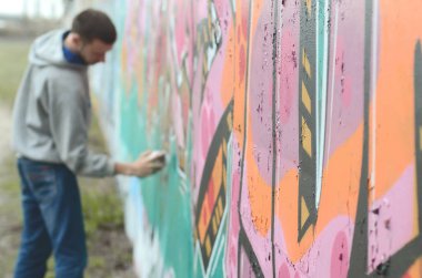 Gri kapüşonlu genç bir adam, yağmurlu havalarda grafiti bir duvar pembe ve yeşil renklerde boyar. Duvar ve bulanık sanatçı parçası odaklanmak