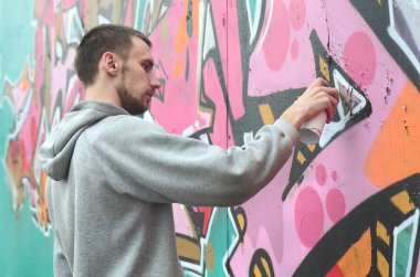 Gri kapüşonlu genç bir adam grafiti bir duvar pembe ve yeşil renklerde yağmurlu havalarda boyar.