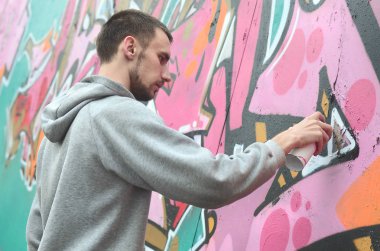 Gri kapüşonlu genç bir adam grafiti bir duvar pembe ve yeşil renklerde yağmurlu havalarda boyar.