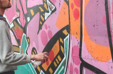 Gri kapüşonlu genç bir adam grafiti bir duvar pembe ve yeşil renklerde yağmurlu havalarda boyar.