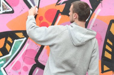 Gri kapüşonlu genç bir adam grafiti bir duvar pembe ve yeşil renklerde yağmurlu havalarda boyar.