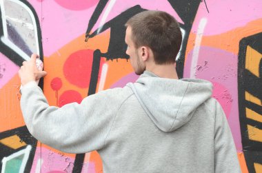 Gri kapüşonlu genç bir adam grafiti bir duvar pembe ve yeşil renklerde yağmurlu havalarda boyar.