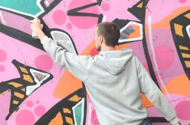 Gri kapüşonlu genç bir adam grafiti bir duvar pembe ve yeşil renklerde yağmurlu havalarda boyar.