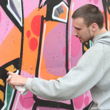 Gri kapüşonlu genç bir adam grafiti bir duvar pembe ve yeşil renklerde yağmurlu havalarda boyar.