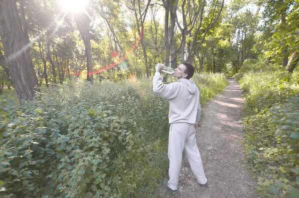 A young guy in a gray sports suit drinks water from a bottle among the trees in the forest. Recreation during a sports run in the open air forest