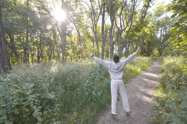 Молодой парень в сером спортивном костюме радуется восходу солнца среди деревьев в лесу. Отдых во время спортивных пробежек в лесу под открытым небом. Восхищение прекрасным рассветом
