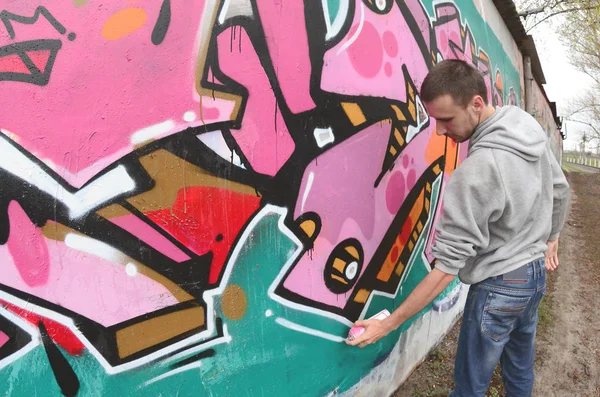 A young guy in a gray hoodie paints graffiti in pink and green colors on a wall in rainy weather. Fisheye shot