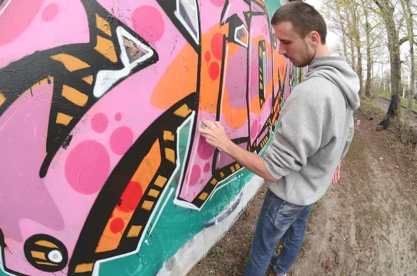 一个穿着灰色帽衫的年轻人在雨天的墙上画着粉红色和绿色的涂鸦 鱼眼射击 — 图库照片