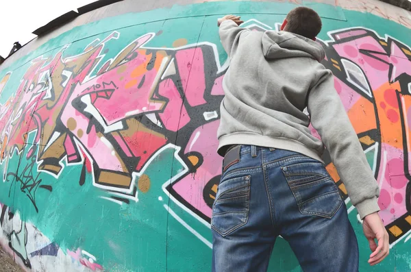 灰色パーカーで若い男は 雨の天気で壁にピンクと緑の色の落書きを描画します 魚眼撮影 — ストック写真