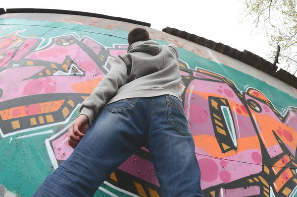 A young guy in a gray hoodie paints graffiti in pink and green colors on a wall in rainy weather. Fisheye shot