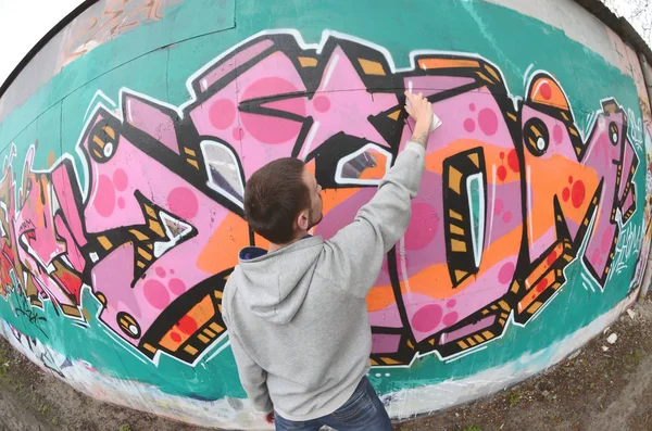 灰色パーカーで若い男は 雨の天気で壁にピンクと緑の色の落書きを描画します 魚眼撮影 — ストック写真