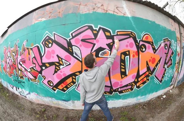 A young guy in a gray hoodie paints graffiti in pink and green colors on a wall in rainy weather. Fisheye shot