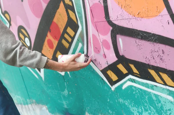 Hand of a young guy in a gray hoodie paints graffiti in pink and green colors on a wall in rainy weather