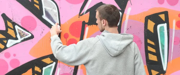 A young guy in a gray hoodie paints graffiti in pink and green colors on a wall in rainy weather