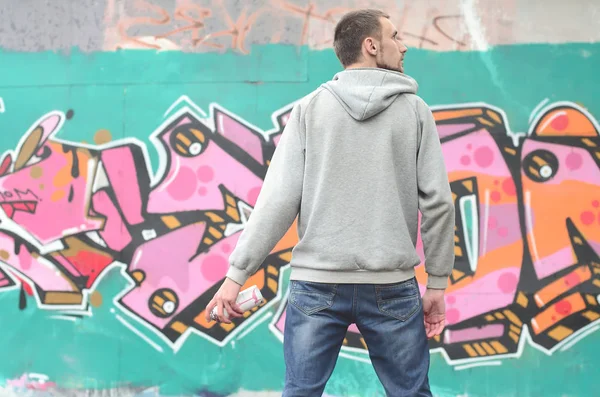 A young graffiti artist in a gray hoodie looks at the wall with his graffiti in pink and green colors on a wall in rainy weather. Street art concept