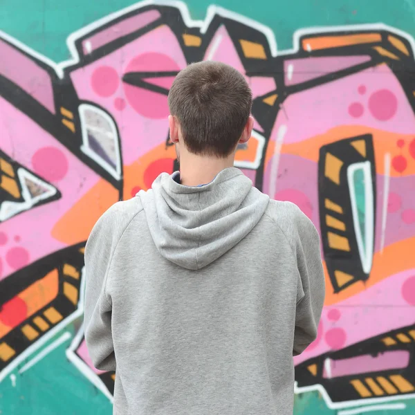 A young graffiti artist in a gray hoodie looks at the wall with his graffiti in pink and green colors on a wall in rainy weather. Street art concept