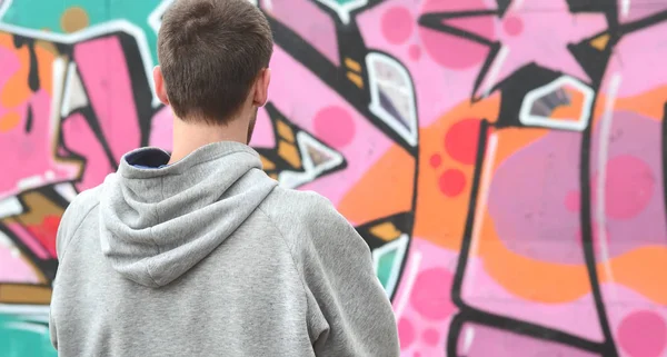 A young graffiti artist in a gray hoodie looks at the wall with his graffiti in pink and green colors on a wall in rainy weather. Street art concept