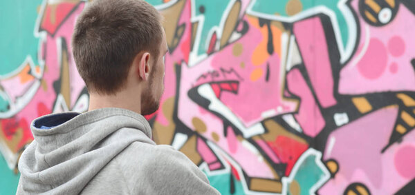 A young graffiti artist in a gray hoodie looks at the wall with his graffiti in pink and green colors on a wall in rainy weather. Street art concept