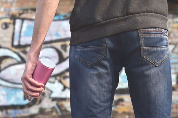 Вид сзади на тело молодого парня в синих грязных джинсах и серый свитер с брызгами краски в руке на фоне старой кирпичной стены, нарисованной граффити
