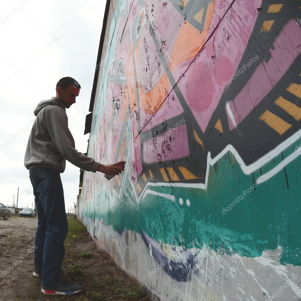 A young guy in a gray hoodie paints graffiti in pink and green colors on a wall in rainy weather. Fisheye shot