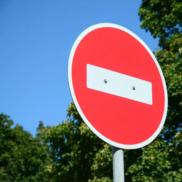 A circular red sign with a white bar indicating NO ENTRY on a grey metal post against a green trees and blue clear sky