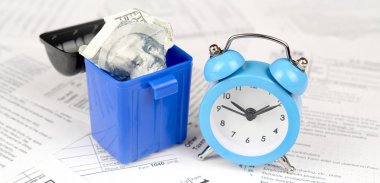 Mavi çalar saat ve çöp kutusu içinde yüz dolarlık banknotu ile birçok Amerikan vergisi boş formlar. Vergiler kavramı ödemek için para kaybı