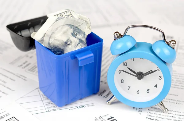 Mavi çalar saat ve çöp kutusu içinde yüz dolarlık banknotu ile birçok Amerikan vergisi boş formlar. Vergiler kavramı ödemek için para kaybı