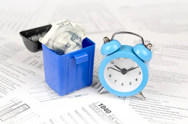 Mavi çalar saat ve çöp kutusu içinde yüz dolarlık banknotu ile birçok Amerikan vergisi boş formlar. Vergiler kavramı ödemek için para kaybı