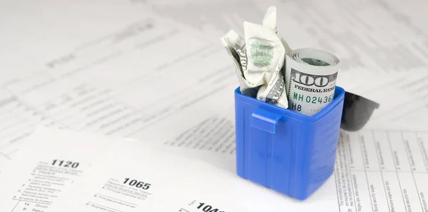 Many american tax blank forms and crumpled hundred dollar bill in trash bin. Loss of money to pay taxes concept