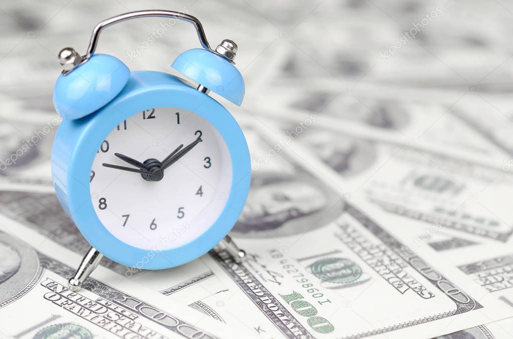 Time management concept. Blue alarm clock lies on a large amount of hundred dollar bills. Time as one of important resources in business