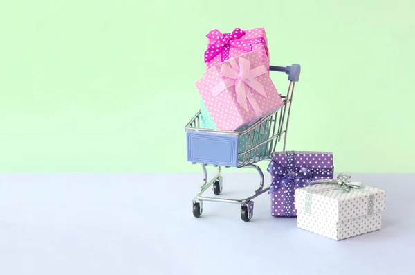 Pequeñas cajas de regalo de diferentes colores con cintas en el carrito de la compra sobre un fondo pastel violeta y azul —  Fotos de Stock